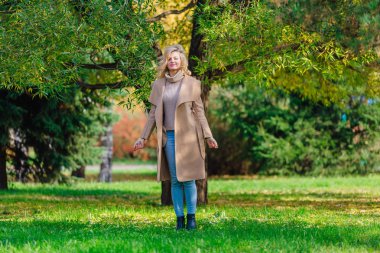 Güzel zarif sarışın kadın ceket ayakta sonbahar park ağaç altında giyinmiş. Güneşli Ekim gün.