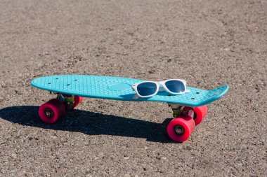 Güneş gözlüğü pembe tekerlekli mavi plastik kaykay kuruş boyutunda bir tahta üzerinde duruyor yolda.