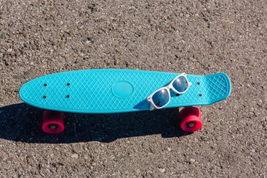 Güneş gözlüğü pembe tekerlekli mavi plastik kaykay kuruş boyutunda bir tahta üzerinde duruyor yolda.