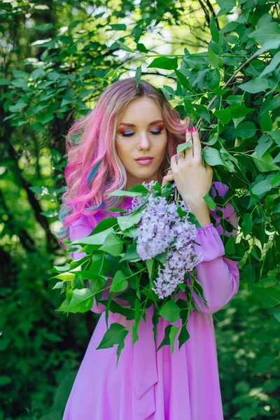 美丽的时尚模特女孩与彩色染发和完美的妆 Standig 旁边的丁香灌木和持有丁香花 — 图库照片