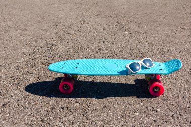 Güneş gözlüğü pembe tekerlekli mavi plastik kaykay kuruş boyutunda bir tahta üzerinde duruyor yolda.
