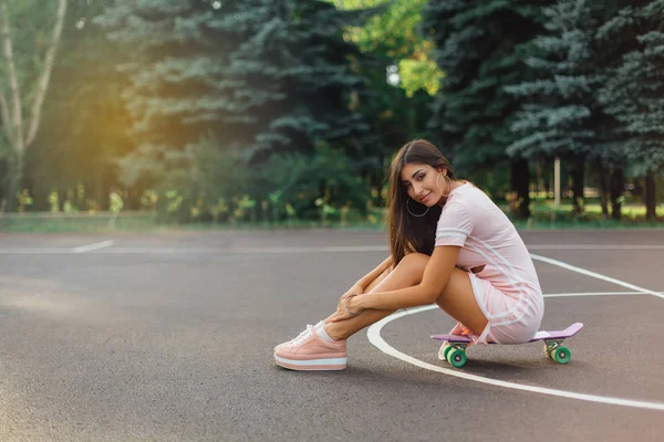 一个微笑迷人的黑发女子坐在篮球场的滑板上的画像 日落时分 穿着时髦的快乐女人正在休息 — 图库照片