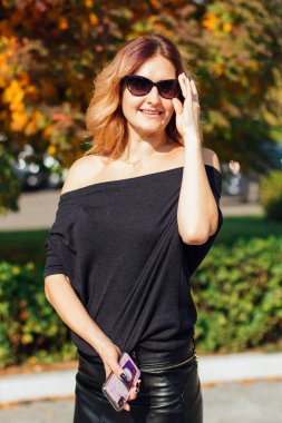 İle siyah elbiseli çekici bir kadın portresi açık omuz ve sonbahar üvez ağacının altında güneş gözlüğü