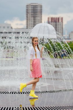 Genç güzel kız sarı çizmeli iki örgüler ve şeffaf şemsiye ile Çeşme duruyor. Şehirde yağmurlu gün.