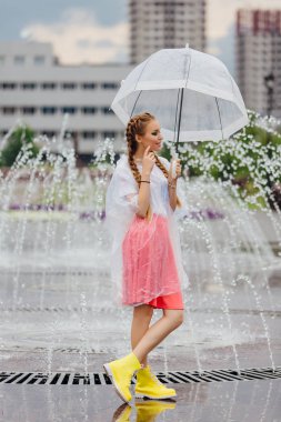 Genç güzel kız sarı çizmeli iki örgüler ve şeffaf şemsiye ile Çeşme duruyor. Şehirde yağmurlu gün.