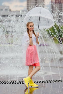 Genç güzel kız sarı çizmeli iki örgüler ve şeffaf şemsiye ile Çeşme duruyor. Şehirde yağmurlu gün.