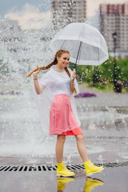 Genç güzel kız sarı çizmeli iki örgüler ve şeffaf şemsiye ile Çeşme duruyor. Şehirde yağmurlu gün.