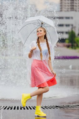 Genç güzel kız sarı çizmeli iki örgüler ve şeffaf şemsiye ile Çeşme duruyor. Şehirde yağmurlu gün.