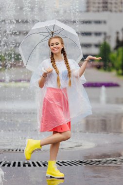 Genç güzel kız sarı çizmeli iki örgüler ve şeffaf şemsiye ile Çeşme duruyor. Şehirde yağmurlu gün.