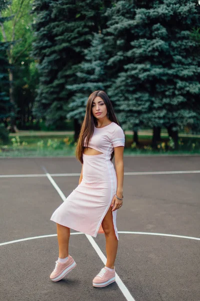 Charming brunette female dressed in a pink dress posing on the basketball court during sunset.