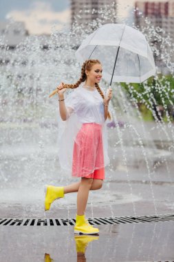 Genç güzel kız sarı çizmeli iki örgüler ve şeffaf şemsiye ile Çeşme duruyor. Şehirde yağmurlu gün.