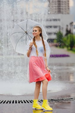 Genç güzel kız sarı çizmeli iki örgüler ve şeffaf şemsiye ile Çeşme duruyor. Şehirde yağmurlu gün.