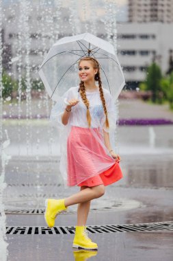 Genç güzel kız sarı çizmeli iki örgüler ve şeffaf şemsiye ile Çeşme duruyor. Şehirde yağmurlu gün.