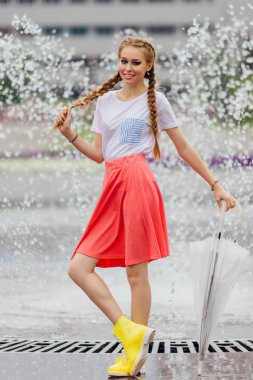 Genç güzel kız sarı çizmeli iki örgüler ve şeffaf şemsiye ile Çeşme duruyor. Şehirde yağmurlu gün.