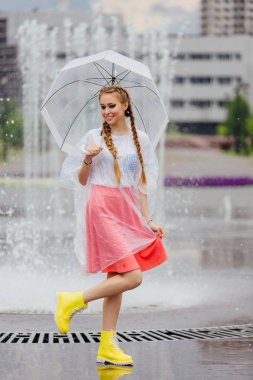 Genç güzel kız sarı çizmeli iki örgüler ve şeffaf şemsiye ile Çeşme duruyor. Şehirde yağmurlu gün.