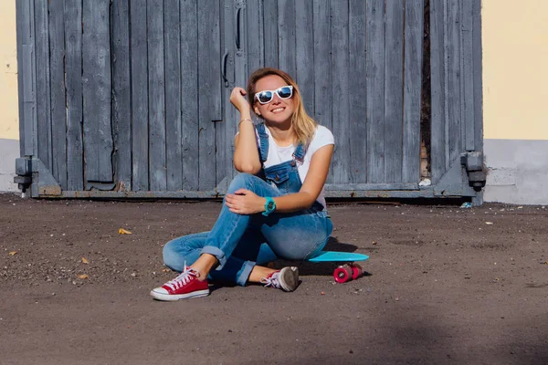 オーバー オールと古い木製の壁の横にある彼女のスケート ボードの上に座ってサングラスを着て笑顔の女性の肖像画 — ストック写真