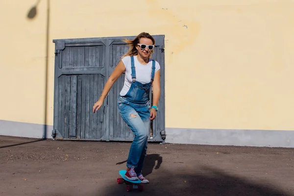 一个微笑的女人的肖像穿着工作服和太阳镜骑在她的塑料滑板旁边的老木墙 — 图库照片