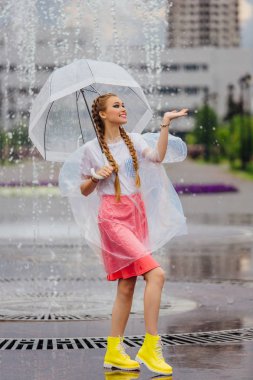 Genç güzel kız sarı çizmeli iki örgüler ve şeffaf şemsiye standları Çeşme yakınındaki.