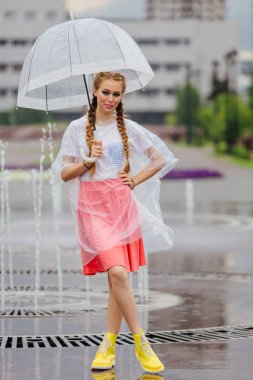 Genç güzel kız sarı çizmeli iki örgüler ve şeffaf şemsiye standları Çeşme yakınındaki.