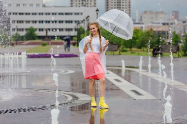 Genç güzel kız sarı çizmeli iki örgüler ve şeffaf şemsiye ile Çeşme duruyor. Şehirde yağmurlu gün.