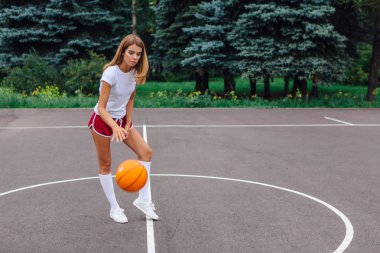 Beyaz tişört, şort ve spor ayakkabı giyen güzel sarışın kız dışarıda basketbol sahasında topla oynuyor. Boşluğu kopyala.