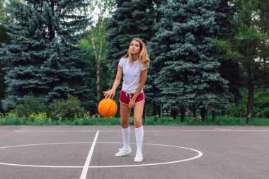 Beyaz tişört, şort ve spor ayakkabı giyen güzel sarışın kız dışarıda basketbol sahasında topla oynuyor. Boşluğu kopyala.
