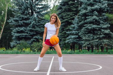 Beyaz tişört, şort ve spor ayakkabı giyen güzel sarışın kız dışarıda basketbol sahasında topla oynuyor. Boşluğu kopyala.