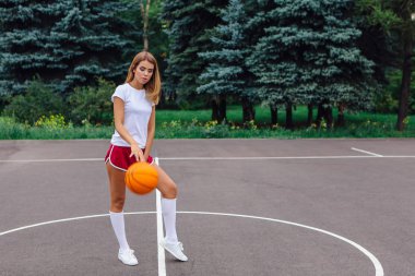 Beyaz tişört, şort ve spor ayakkabı giyen güzel sarışın kız dışarıda basketbol sahasında topla oynuyor. Boşluğu kopyala.