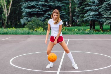 Beyaz tişört, şort ve spor ayakkabı giyen güzel sarışın kız dışarıda basketbol sahasında topla oynuyor. Boşluğu kopyala.