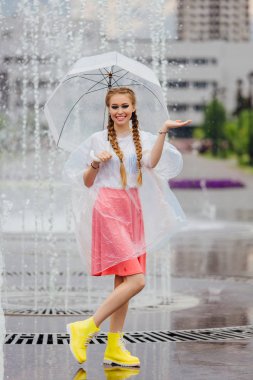 Genç güzel kız sarı çizmeli iki örgüler ve şeffaf şemsiye ile Çeşme duruyor. Şehirde yağmurlu gün.