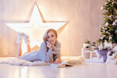Güzel genç sarışın kadın döşeme ve bir tatil iç Noel ağacı altında kitap okuma beyaz kazak giymiş