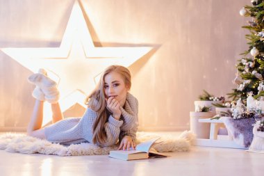 Güzel genç sarışın kadın döşeme ve bir tatil iç Noel ağacı altında kitap okuma beyaz kazak giymiş