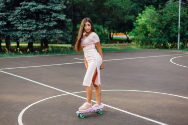 Bir gülümseyen şirin esmer kadın ayakta onu kaykay üzerinde bir basketbol sahası üzerinde portresi. Mutlu kadın trendy bakışla günbatımı sırasında mola.