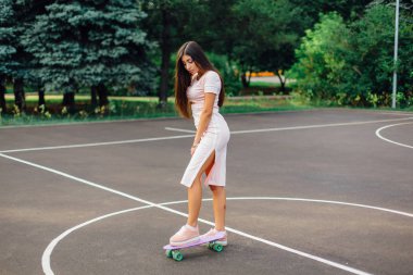 Bir gülümseyen şirin esmer kadın ayakta onu kaykay üzerinde bir basketbol sahası üzerinde portresi. Mutlu kadın trendy bakışla günbatımı sırasında mola.