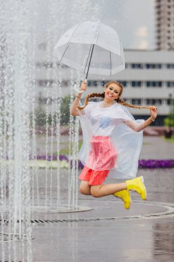 Genç güzel kız sarı çizmeli iki örgüler ve şeffaf şemsiye Çeşme atlama ile. Şehirde yağmurlu gün.