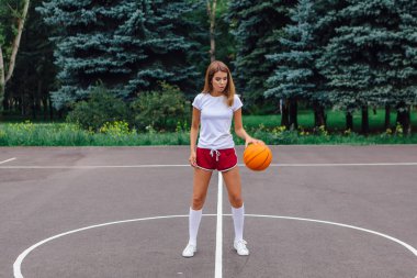 Beyaz tişört, şort ve spor ayakkabı giyen güzel sarışın kız dışarıda basketbol sahasında topla oynuyor. Boşluğu kopyala.