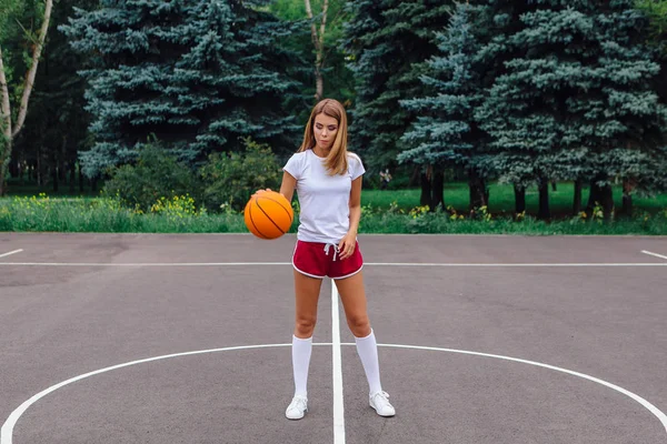 Beyaz tişört, şort ve spor ayakkabı giyen güzel sarışın kız dışarıda basketbol sahasında topla oynuyor. Boşluğu kopyala.