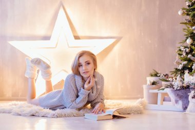 Güzel genç sarışın kadın döşeme ve bir tatil iç Noel ağacı altında kitap okuma beyaz kazak giymiş