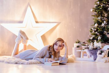 Güzel genç sarışın kadın döşeme ve bir tatil iç Noel ağacı altında kitap okuma beyaz kazak giymiş