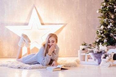 Güzel genç sarışın kadın döşeme ve bir tatil iç Noel ağacı altında kitap okuma beyaz kazak giymiş