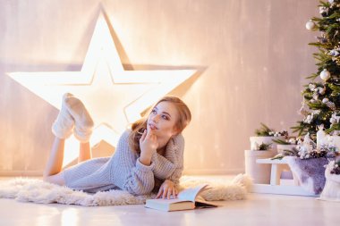 Güzel genç sarışın kadın döşeme ve bir tatil iç Noel ağacı altında kitap okuma beyaz kazak giymiş
