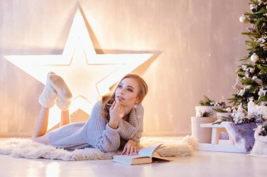 Güzel genç sarışın kadın döşeme ve bir tatil iç Noel ağacı altında kitap okuma beyaz kazak giymiş