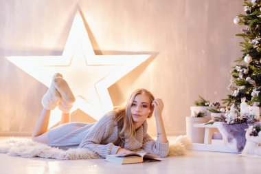 Güzel genç sarışın kadın döşeme ve bir tatil iç Noel ağacı altında kitap okuma beyaz kazak giymiş