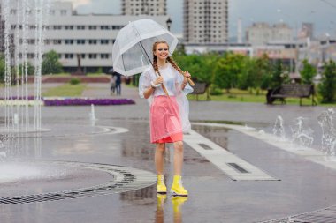 Genç güzel kız sarı çizmeli iki örgüler ve şeffaf şemsiye ile Çeşme duruyor. Şehirde yağmurlu gün.