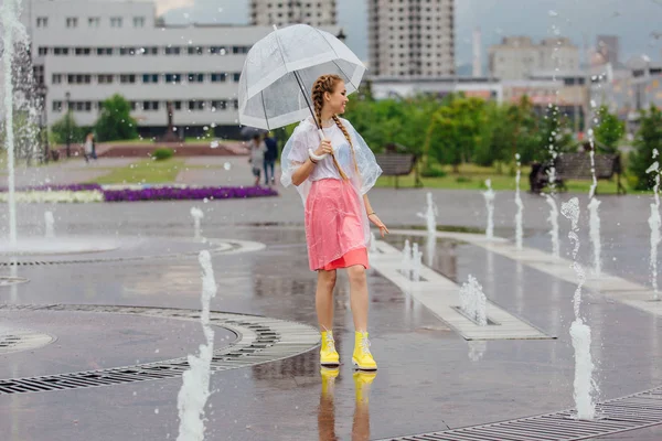 透明傘と黄色のブーツでお下げ髪若いきれいな女の子は噴水のそばに立ちます 雨の日の市 — ストック写真