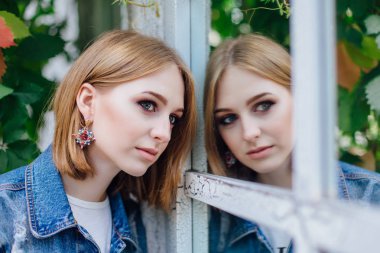 Moda kız waching yansıması, büyük boy kot ceket ve güzel kulak halkaları, ayna pencere ayakta.