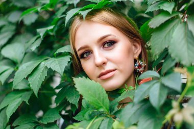 Makyaj olan güzel bir kadın portresi ve kulak halkaları, üzüm yapraklarda gizleme.