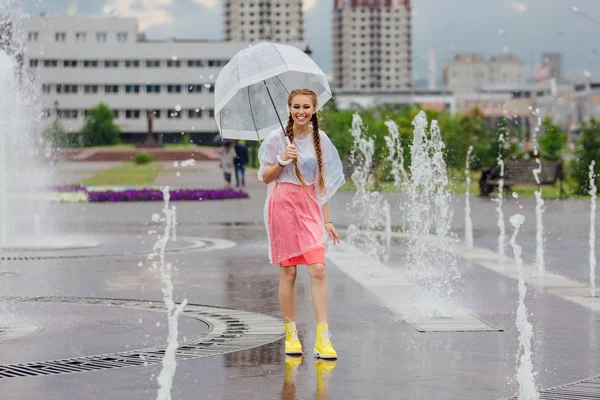 Gadis cantik muda dengan dua kepang sepatu bot kuning dan dengan payung transparan berdiri di dekat air mancur . — Stok Foto