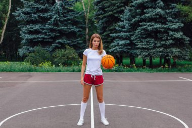 Güzel genç kız beyaz tişört, şort ve spor ayakkabısı, giyinmiş bir basketbol sahası üzerinde bir top ile çalış.