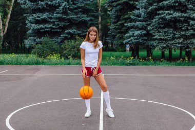 Güzel genç kız beyaz tişört, şort ve spor ayakkabısı, giyinmiş bir basketbol sahası üzerinde bir top ile çalış.